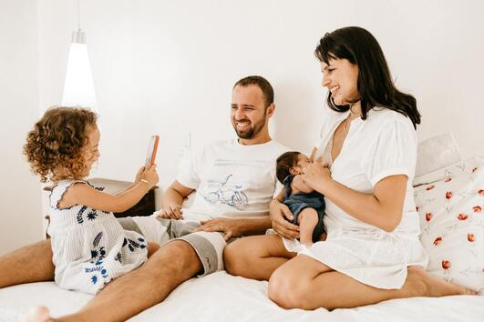 A family sitting together in bed, a newborn nursing from the mother while a small child sits with her father. The small child is taking a photo of her family